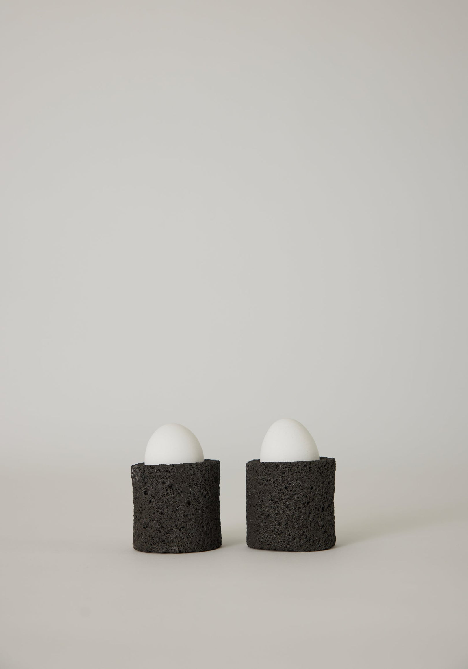 Egg Cups: Two white eggs in handcrafted black holders made from lava stone, showcasing a unique porous texture, designed by artist Louis Crombag.