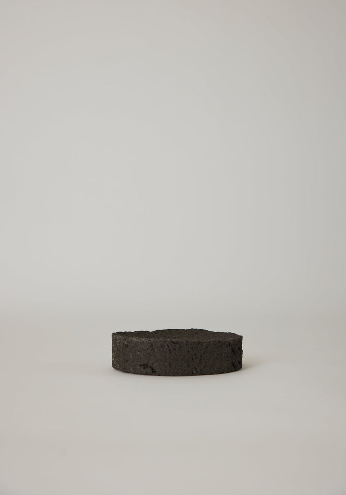 Round Tray crafted from leftover lava stone, appears as a porous black disc. Handcrafted by Louis Crombag, each unique piece measures 2 cm high and 13.5 cm wide.