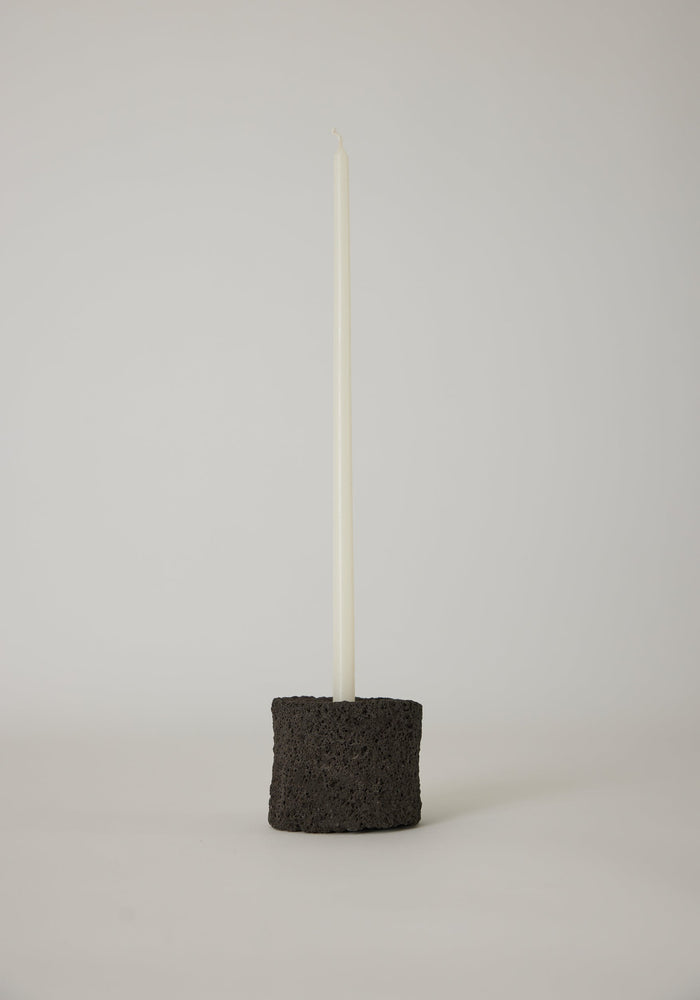 Round Candle Holder: Slim white taper candle in a handcrafted black lava stone holder, showcasing minimalist design and artisanal craftsmanship by Dutch artist Louis Crombag.