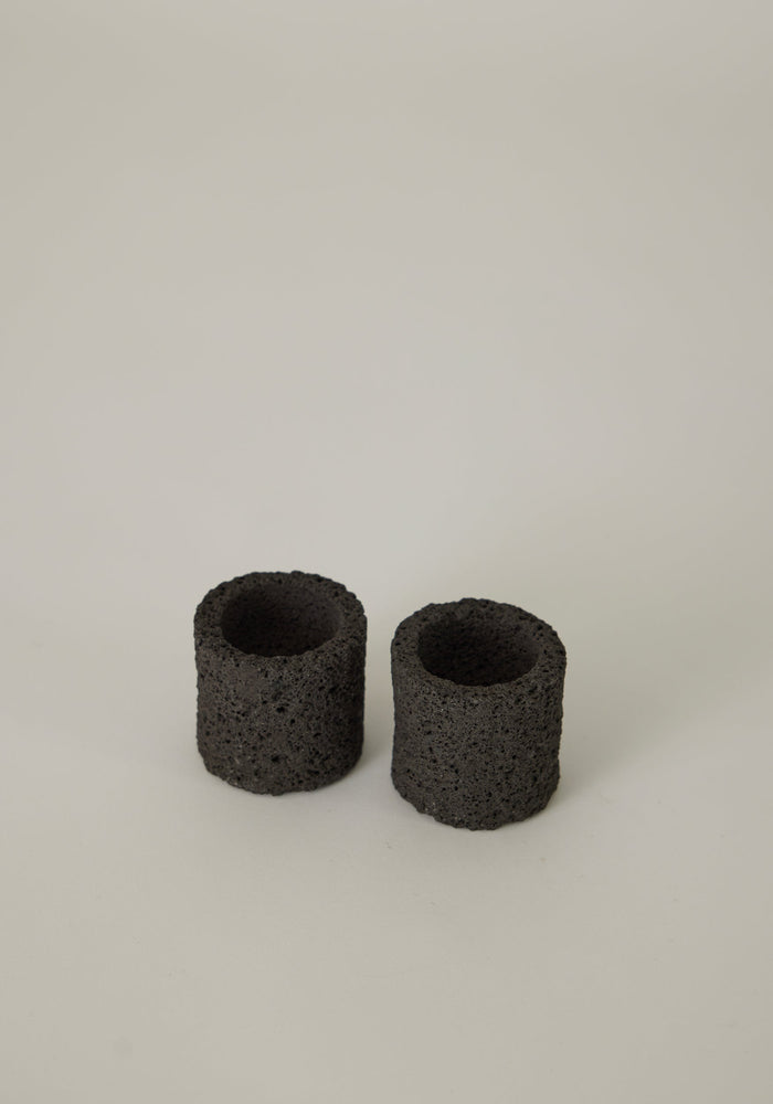 Egg Cups: Two handcrafted, cylindrical containers made from porous lava stone by Dutch artist Louis Crombag, showcasing unique textures on a smooth surface.
