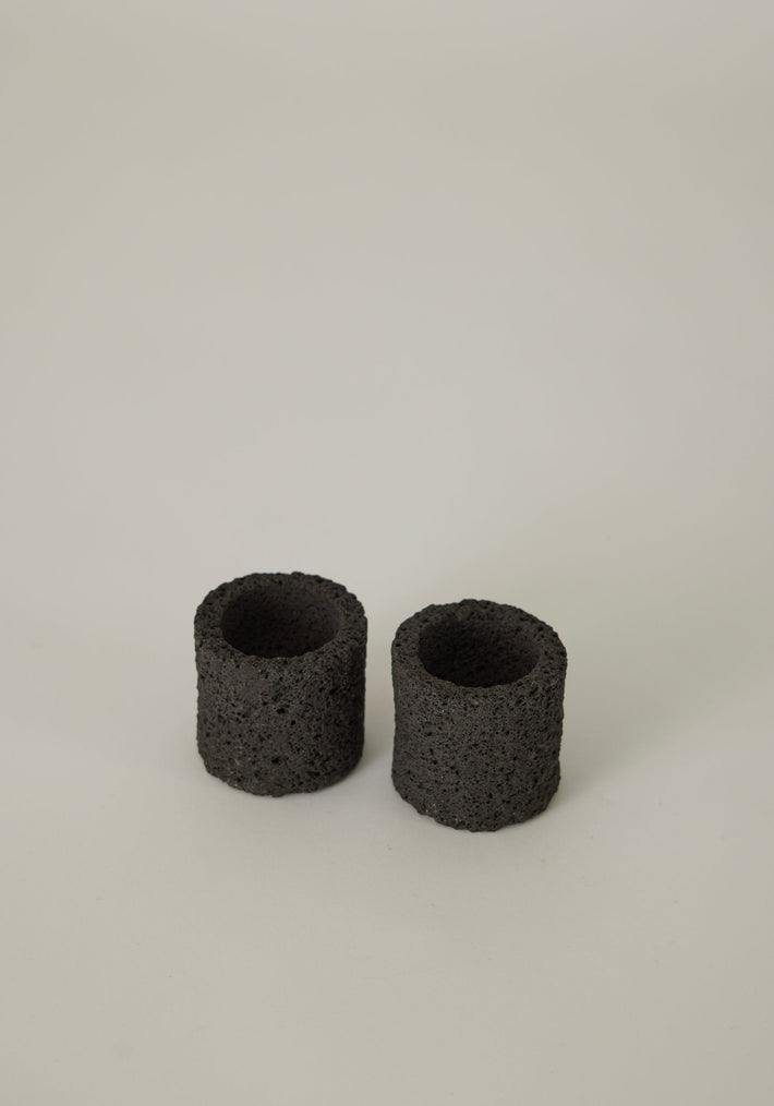 Egg Cups: Two handcrafted, cylindrical containers made from porous lava stone by Dutch artist Louis Crombag, showcasing unique textures on a smooth surface.