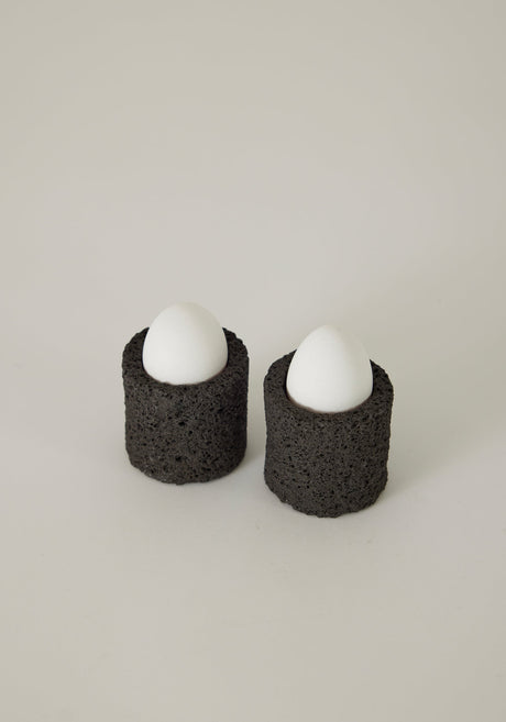 Egg Cups: Two handcrafted lava stone cups holding eggs, set on a linen napkin with a decorative teaspoon, showcasing unique craftsmanship by Dutch artist Louis Crombag.