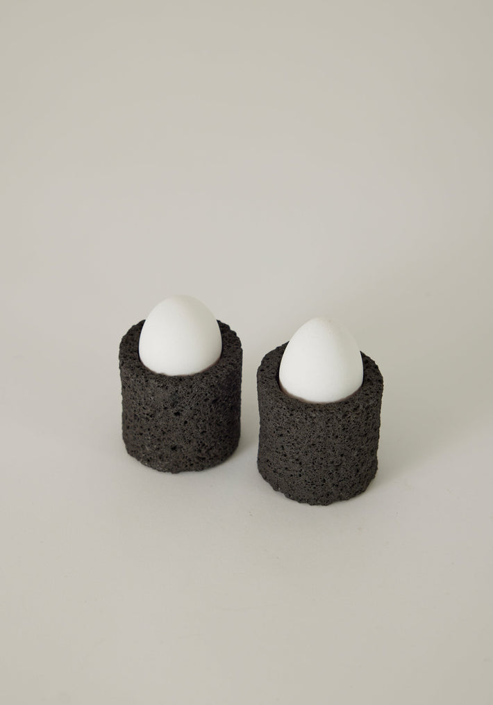 Egg Cups: Two white egg shells sit in rough-textured, black lava stone cylinders, handcrafted by Dutch artist Louis Crombag, highlighting their unique, artisanal nature.