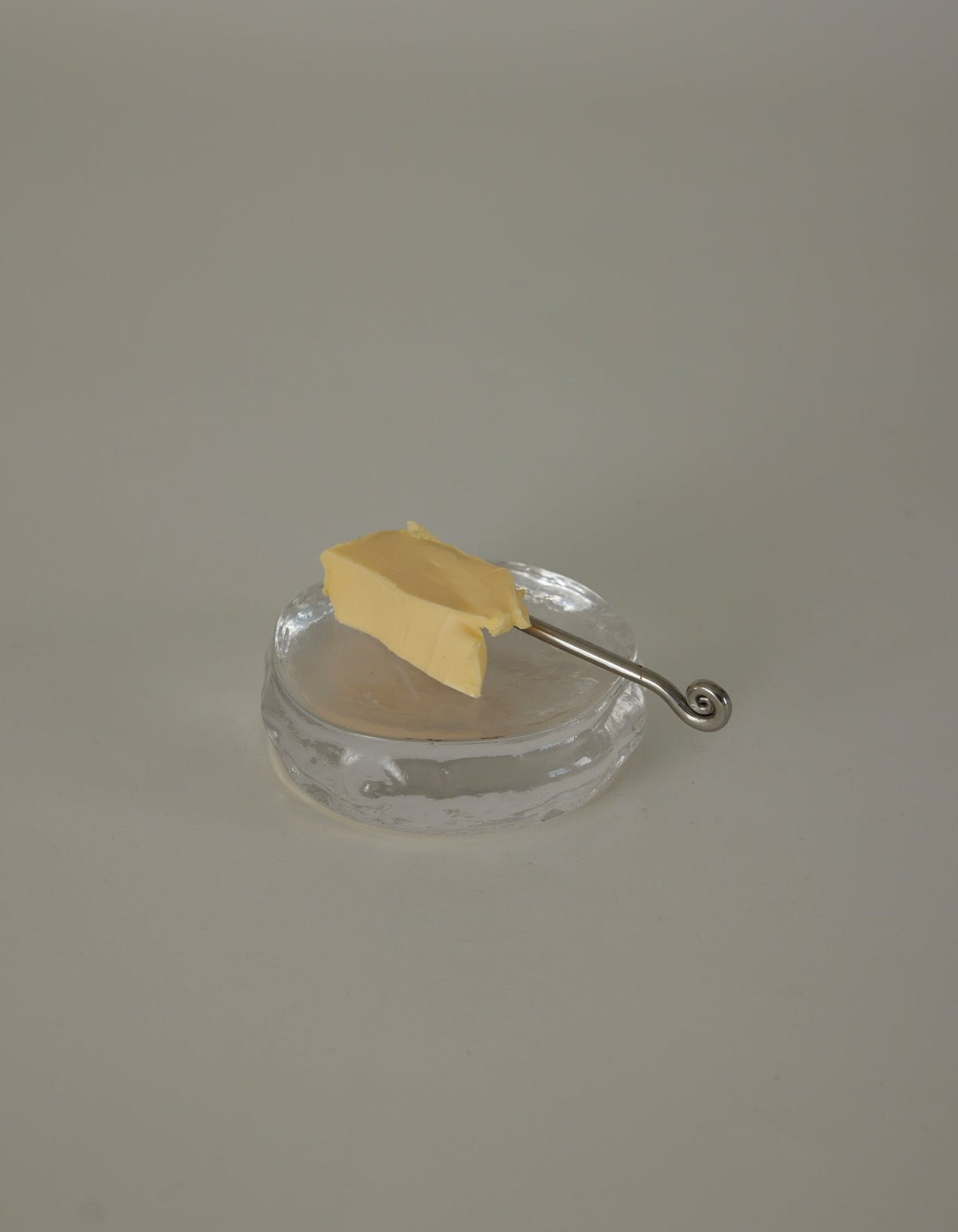 Butter plate | Glass by Szkło Studio: An irregular clear glass dish with a silver spreader knife and butter pat, showcasing unique, hand-blown craftsmanship.