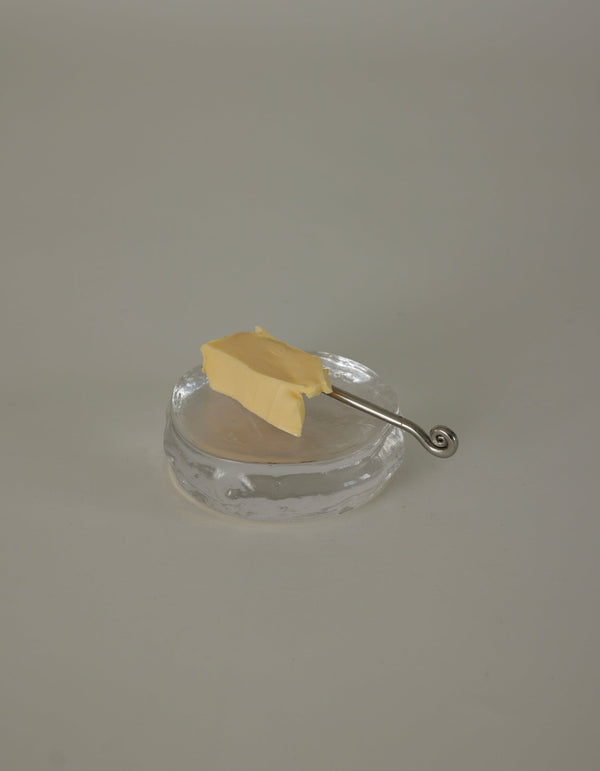 Butter plate | Glass by Szkło Studio: An irregular clear glass dish with a silver spreader knife and butter pat, showcasing unique, hand-blown craftsmanship.