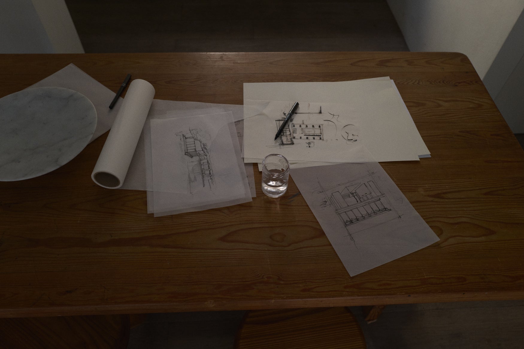Table with architectural drawings, a glass of water, and a pen on a wooden surface.