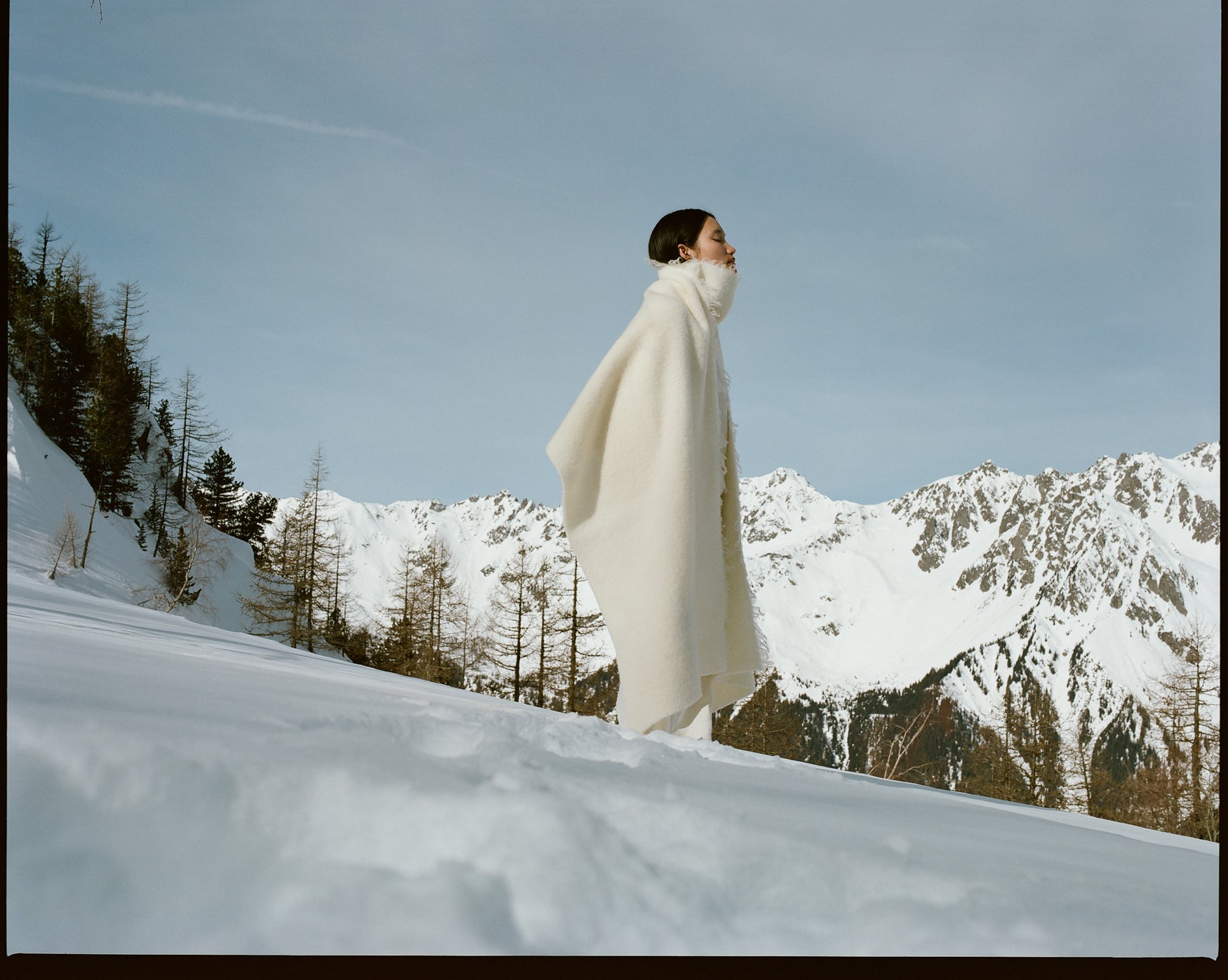 Woman standing in a cream wool poncho on a snowy hill with mountains and a blue sky in the background