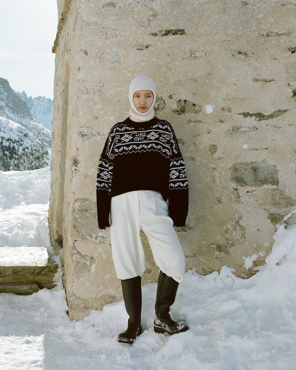 Woman leaning against a rustig stone wall wearing a knitted balaclava and fair isle jumper with a snow and a mountain on the background