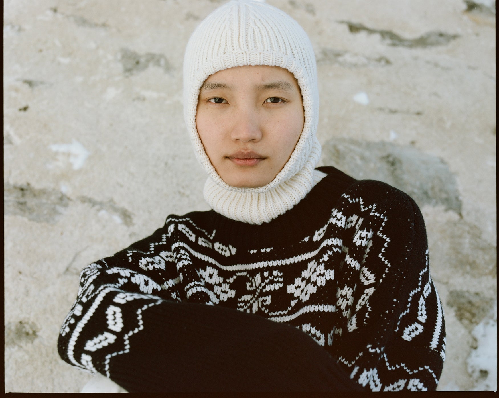 Portrait of a woman wearing a cream knitted balaclava and a black and white fair isle sweater against a rustic stone wall background