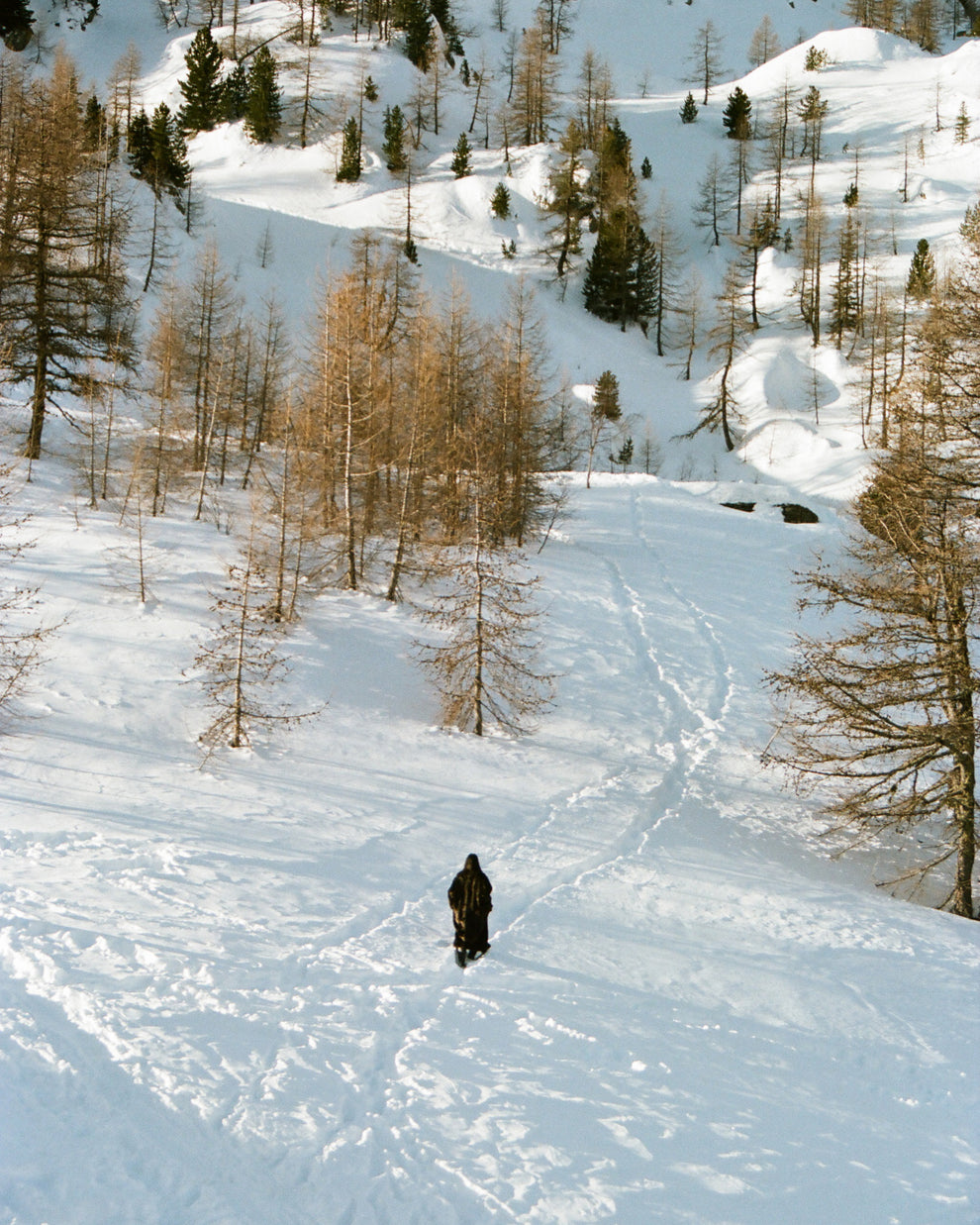 Woman from afar walking in a snowy mountain landscape wearing a long faux fur dark brown coat