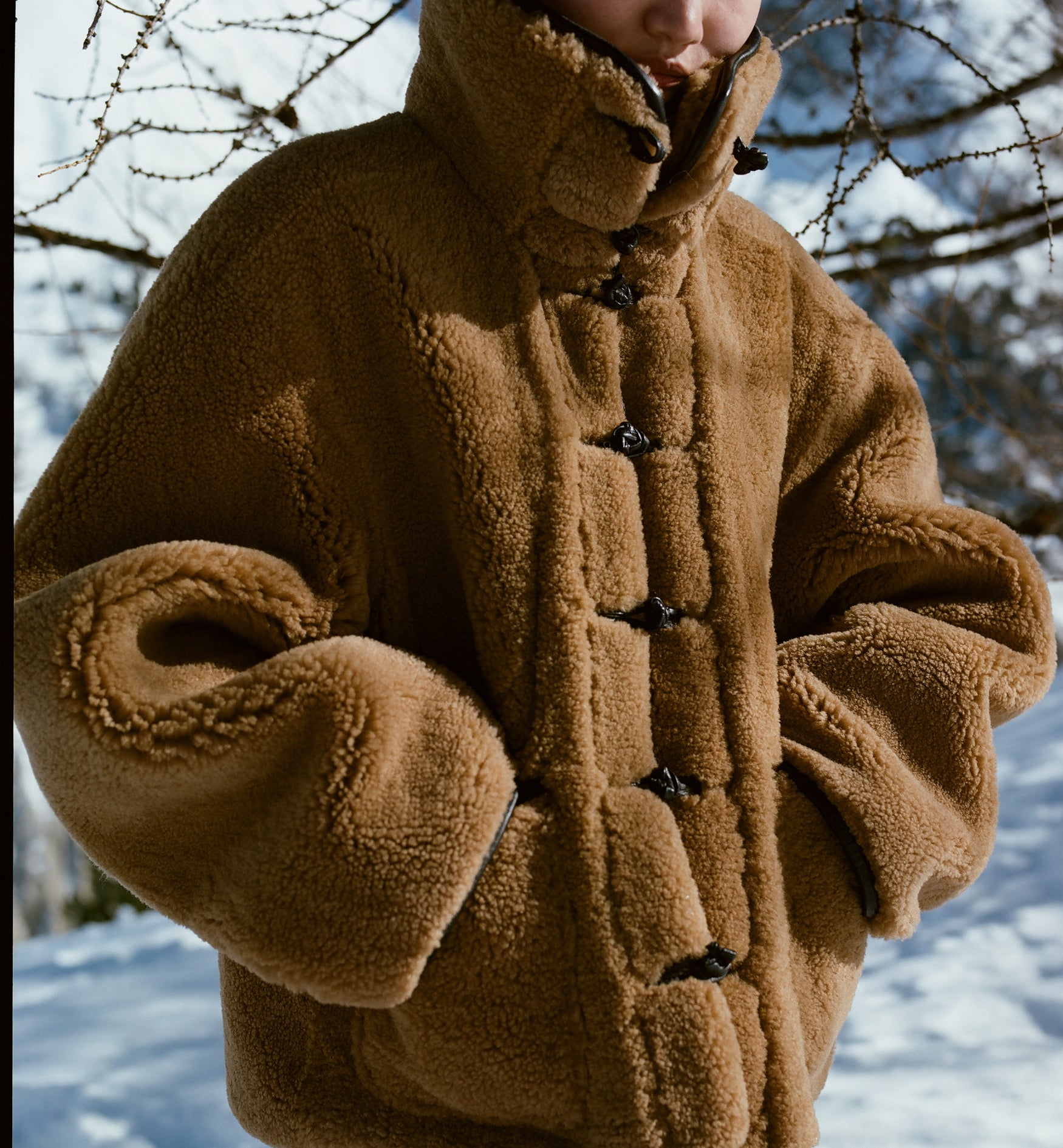 Close up image of a cognac colored shearling jacket with a high funnel neck and snow in the background