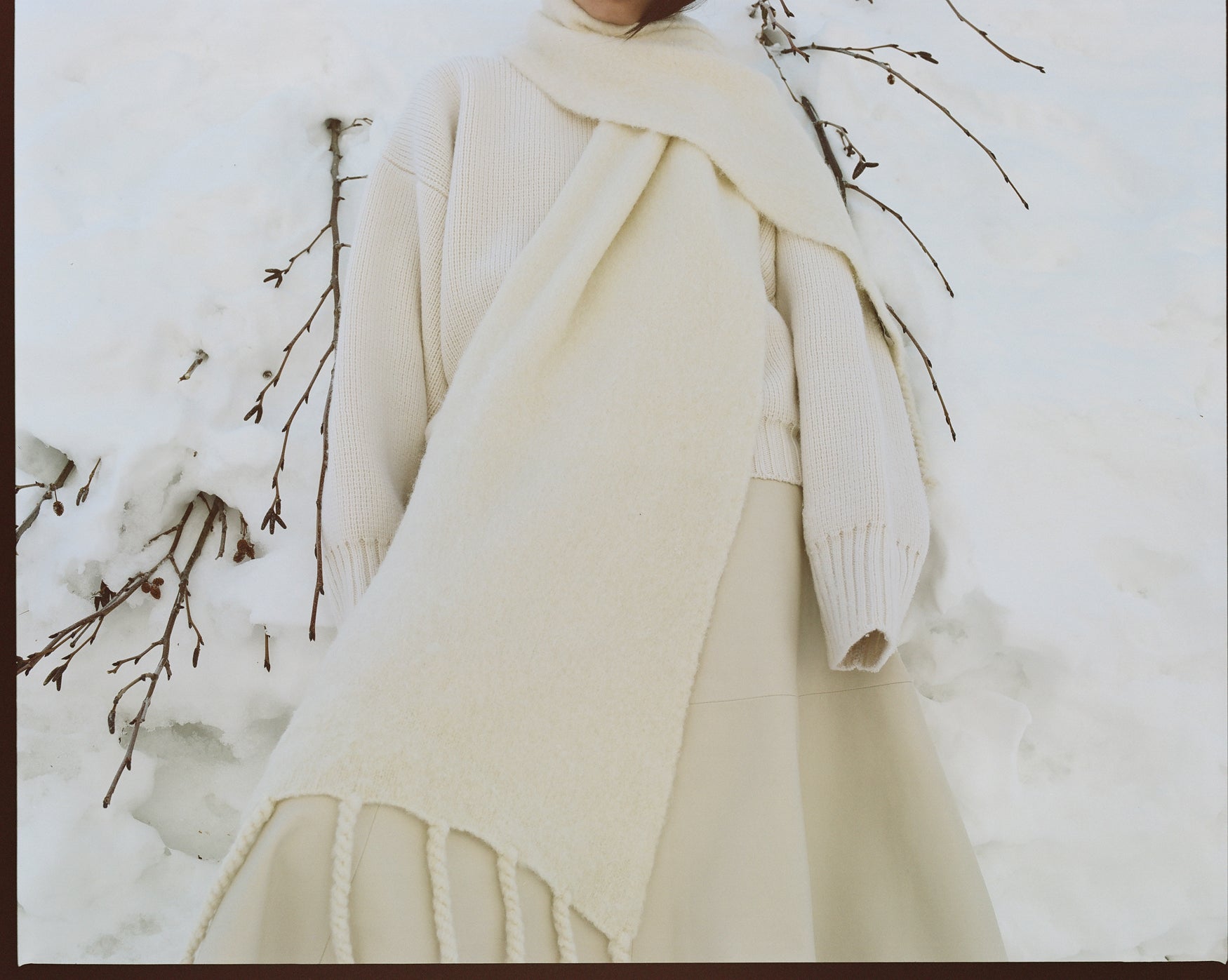Woman leaning against a snow wall wearing a cream knitted sweater, cream knitted scarf, cream leather a-line skirt