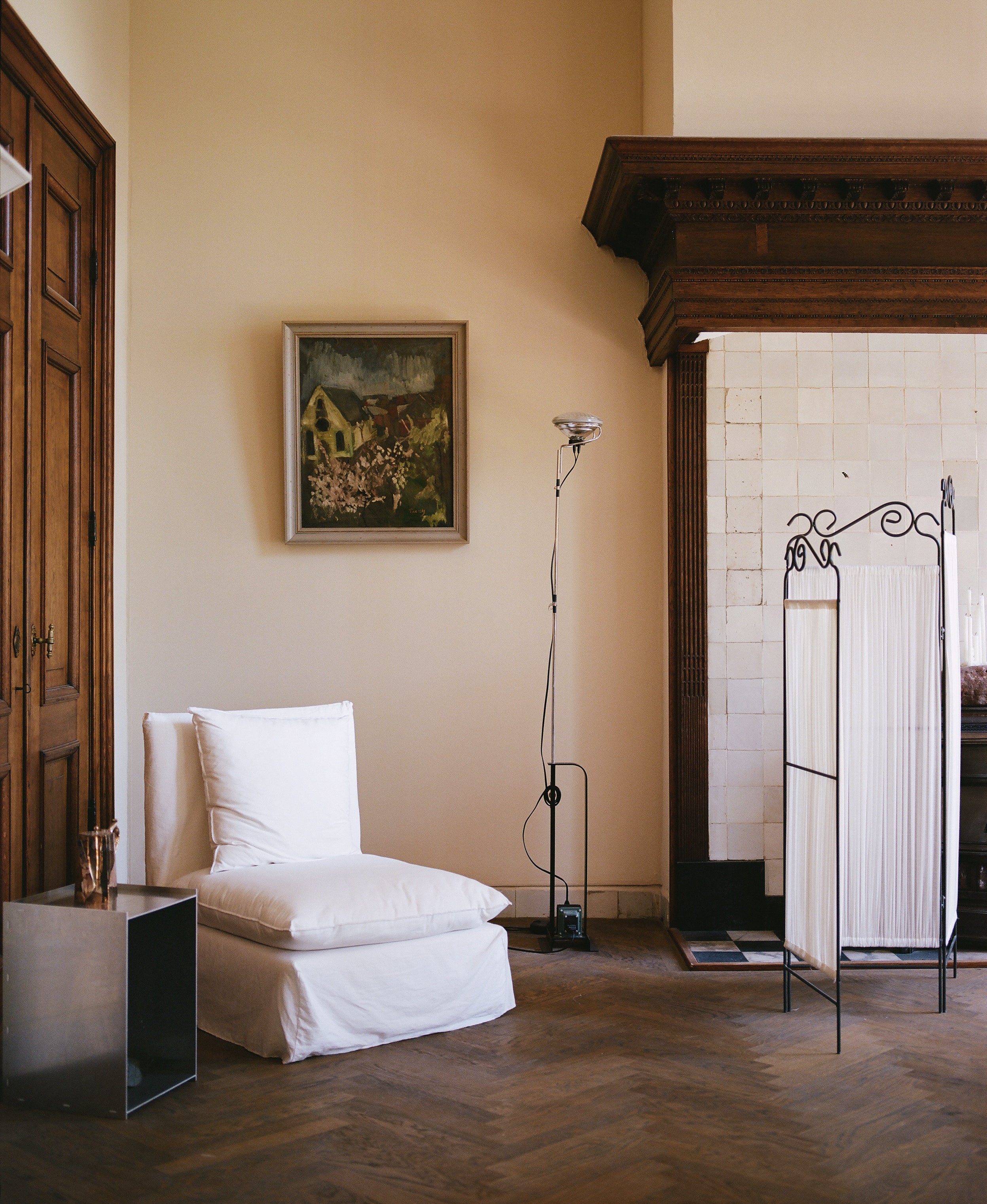 White fabric covered chair in a 17th century room with a tiled fire place, decorative room divider and stainless steel side table