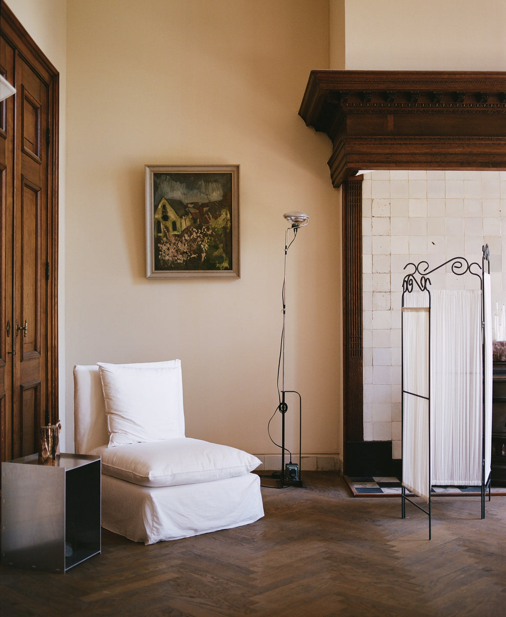 White fabric covered chair in a 17th century room with a tiled fire place, decorative room divider and stainless steel side table