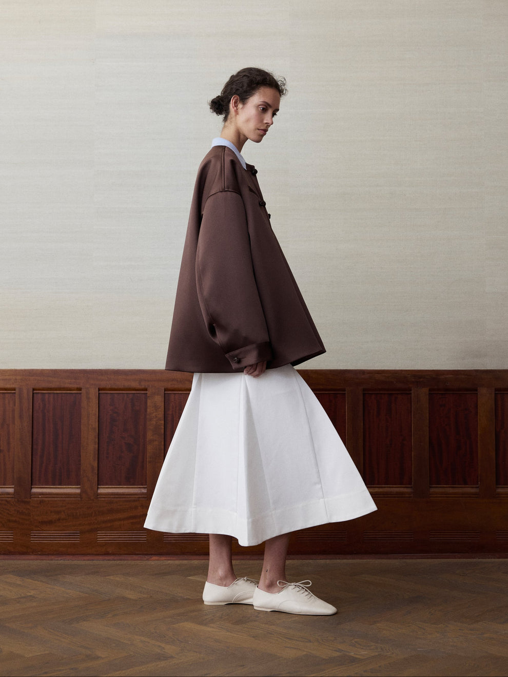 Woman wearing an brown colored heavy satin jacket and a white sculptural skirt against a linen and wood paneled background 