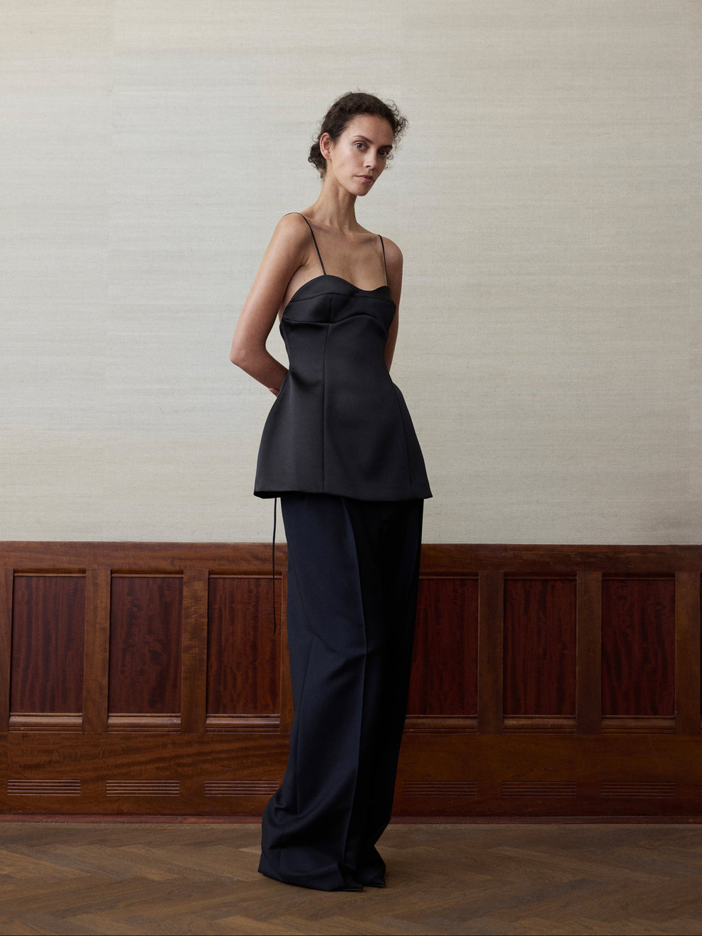 Woman wearing a heavy satin black spaghetti strap bustier top and navy trousers against a linen and wood paneled background 