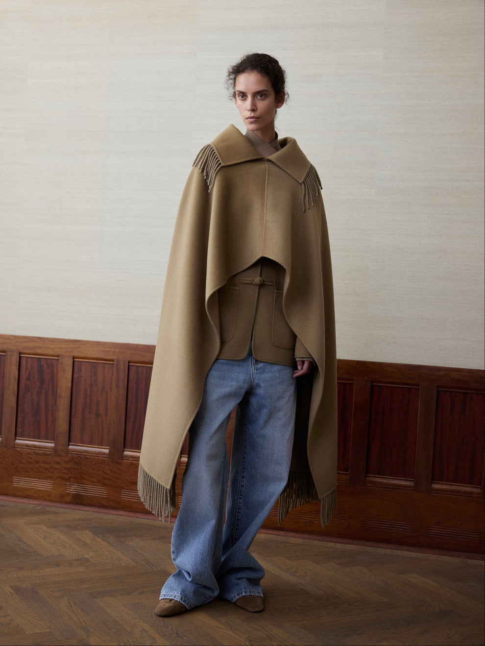 Woman wearing a camel colored fringed poncho and blue jeans against a linen and wood paneled background