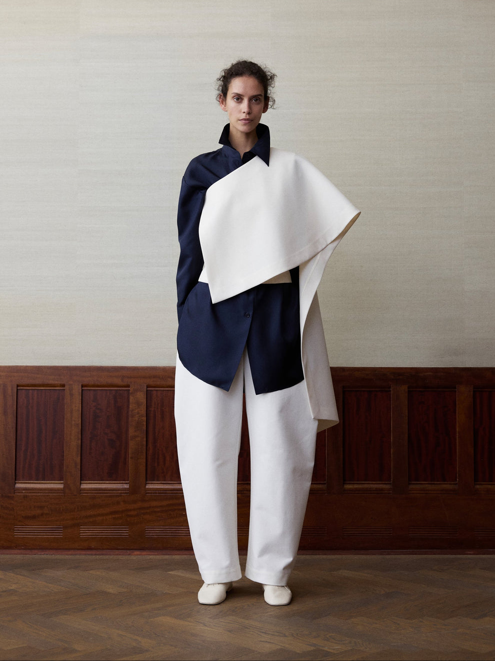 Woman wearing a navy silk shirt, white a symmetrical cape top and white balloon trousers against a linen and wood paneled background 