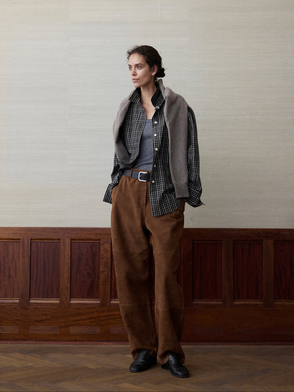 Woman wearing a check shirt and suede trousers against a linen and wood paneled background 