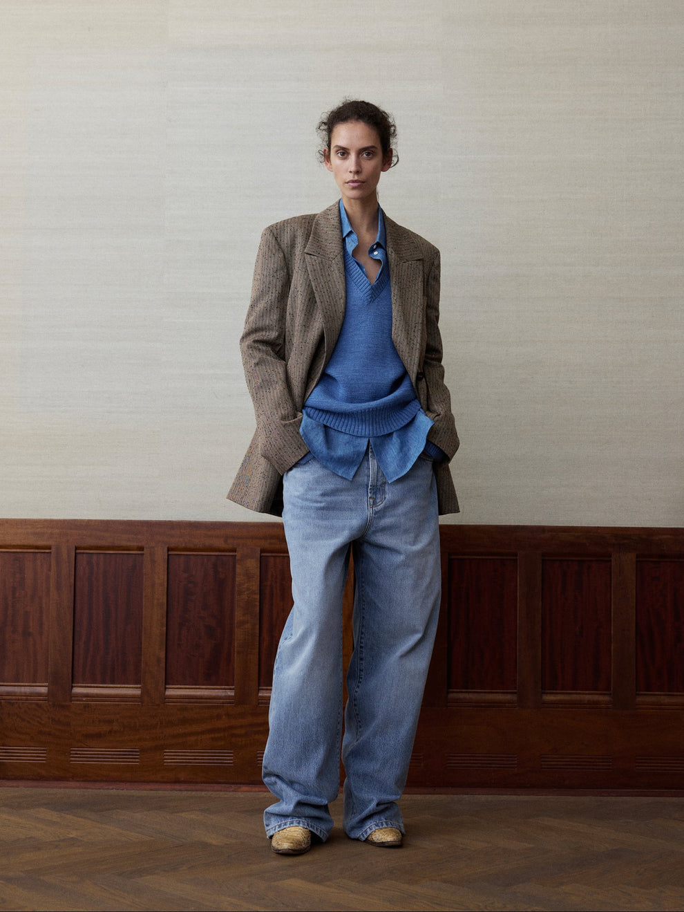 Woman wearing a blue v-neck knitted sweater, blue jeans and a slub melange brown blazer against a linen and wood paneled background