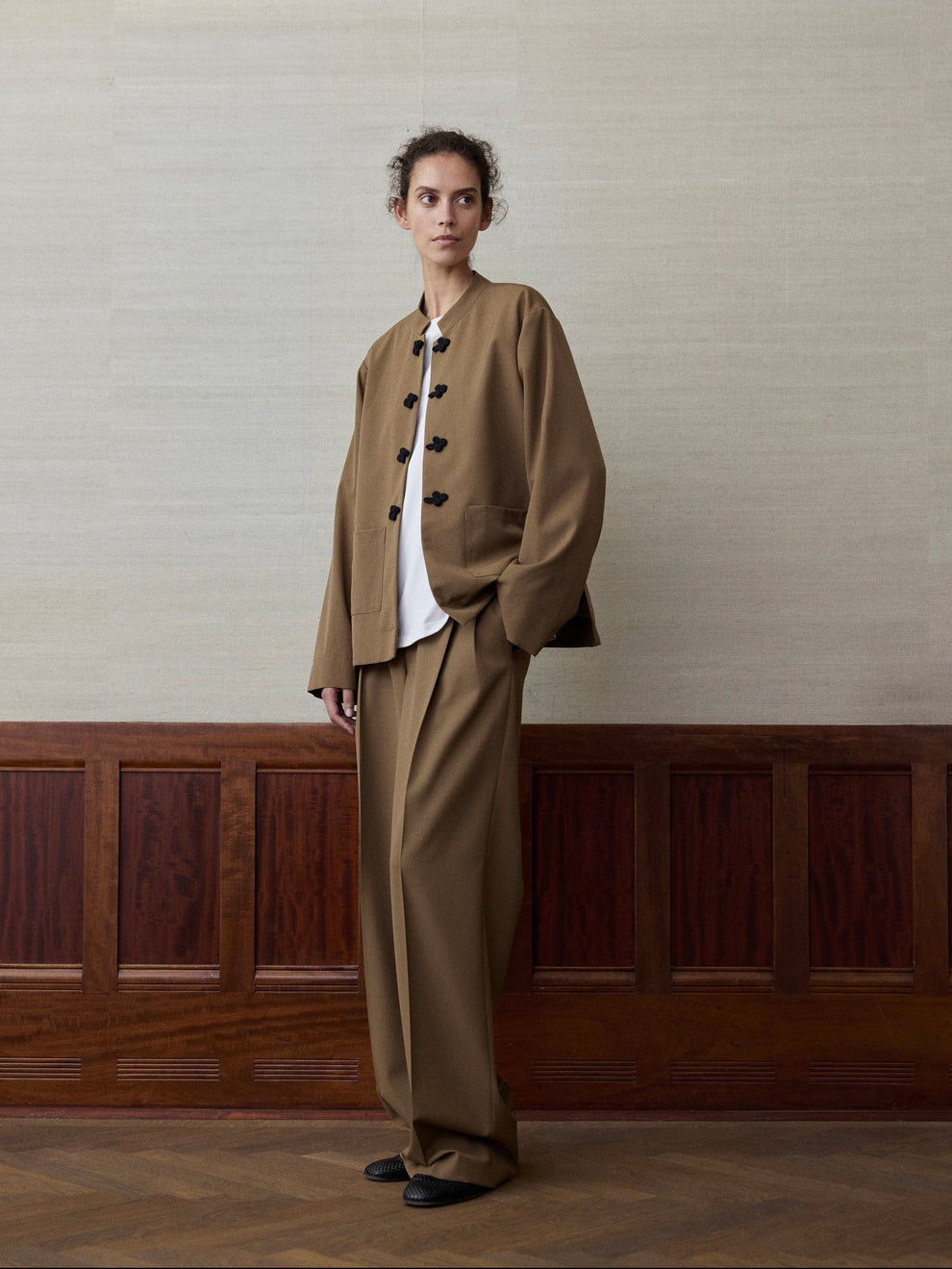 Woman wearing a mocha colored suit consisting of a jacket with mandarin closure and matching trousers against a linen and wood paneled background