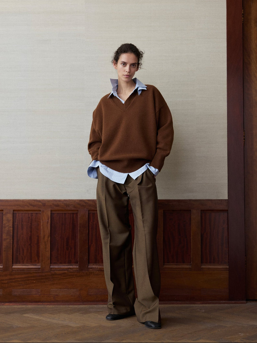 Woman wearing a rust colored v-neck knitted sweater and golden brown colored trousers against a linen and wood paneled background