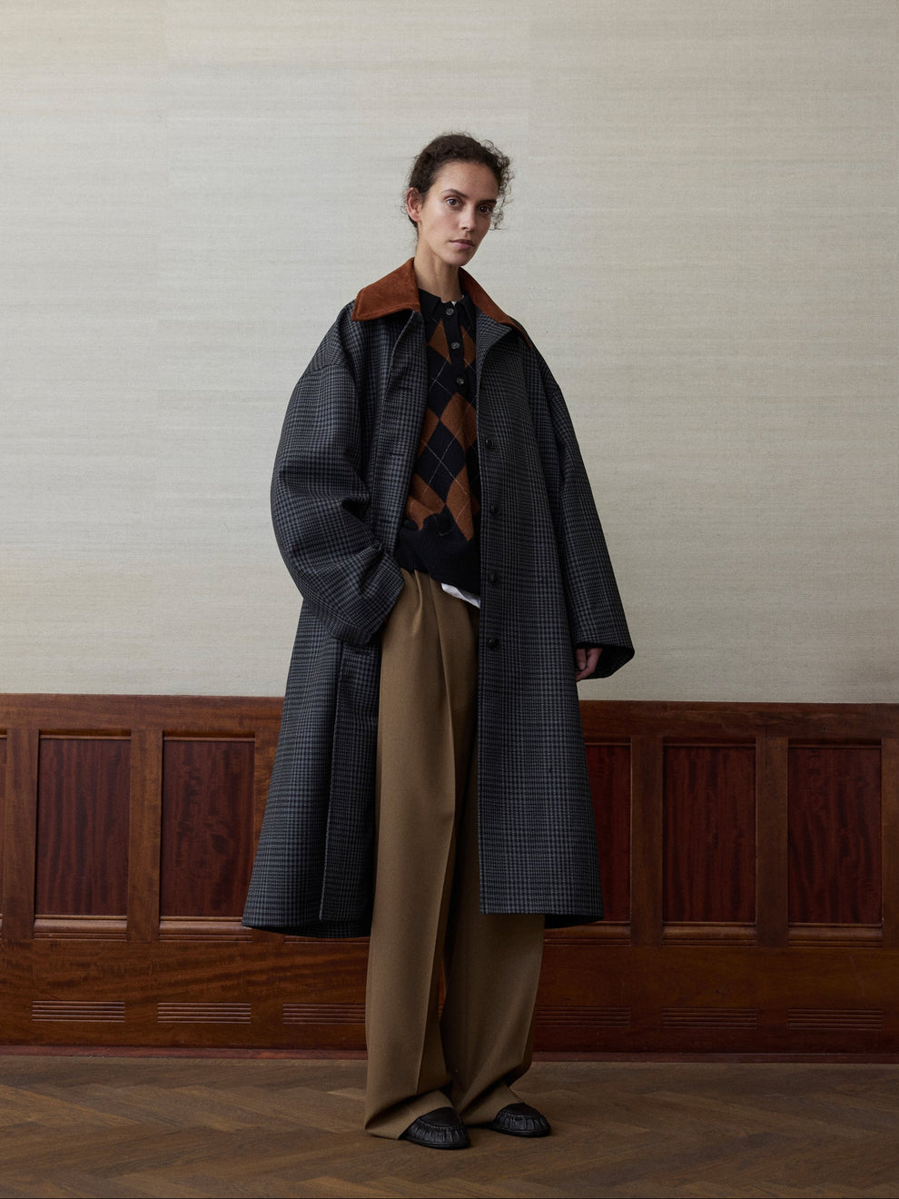 Woman wearing a black and grey check coat with suede collar and golden brown trousers against a linen and wood paneled background