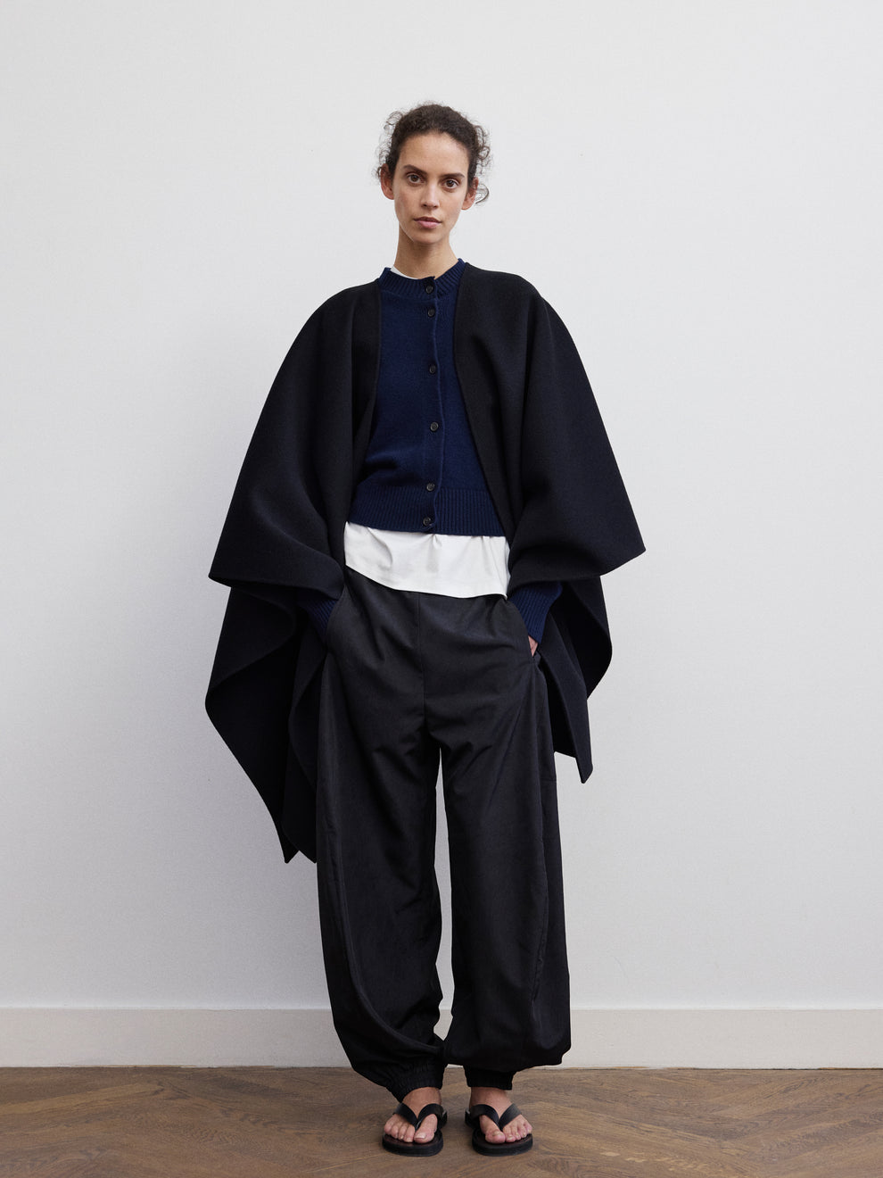 Woman wearing a white t-shirt, navy button wool cardigan, black balloon track pants and black poncho on top against a white background