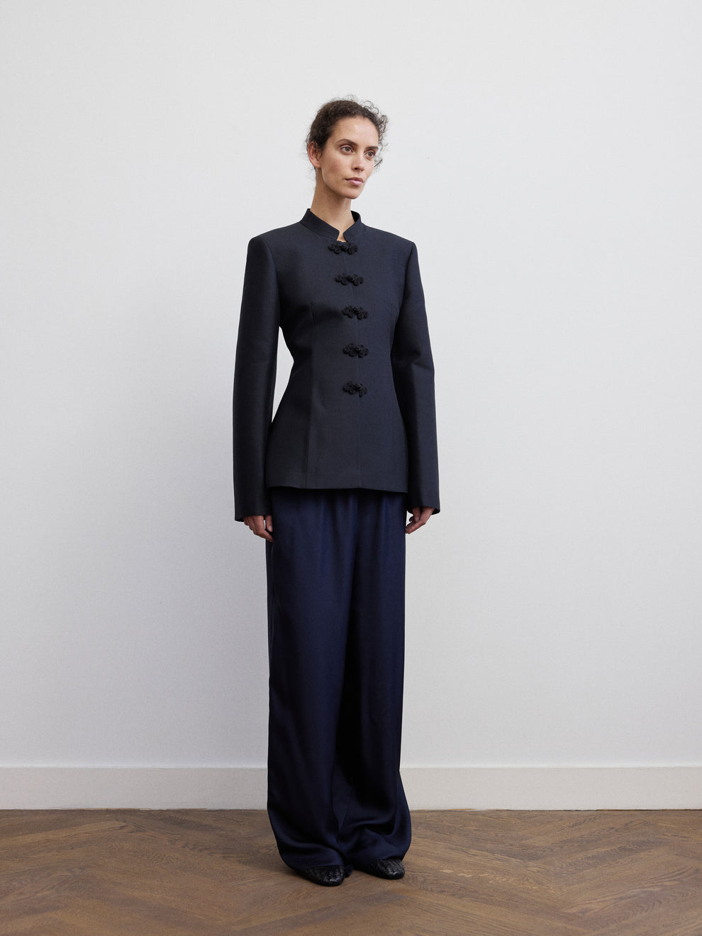 Woman wearing a black blazer with mandarin closure and navy silk pants against a white background 
