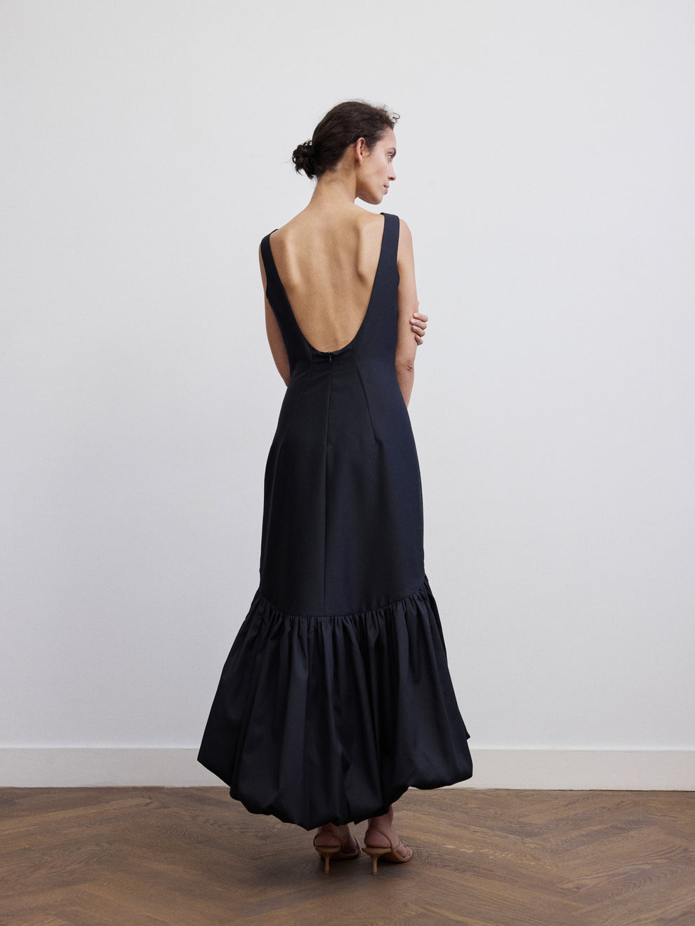 Woman photographed from the back wearing a long raw silk black dress with low back and voluminous bottom against a white background