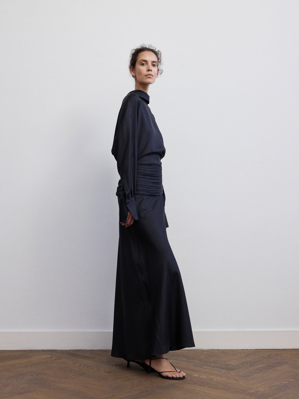 Woman wearing a long silk black dress with rushed detail at the waist against white background