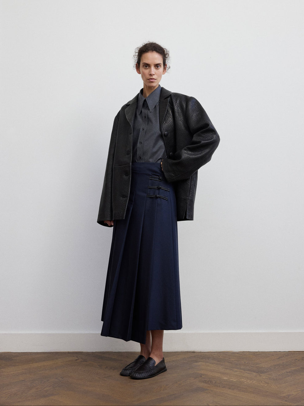 Woman wearing a grey flanel fitted shirt, navy pleated skirt and black grained leather blazer against a white background 