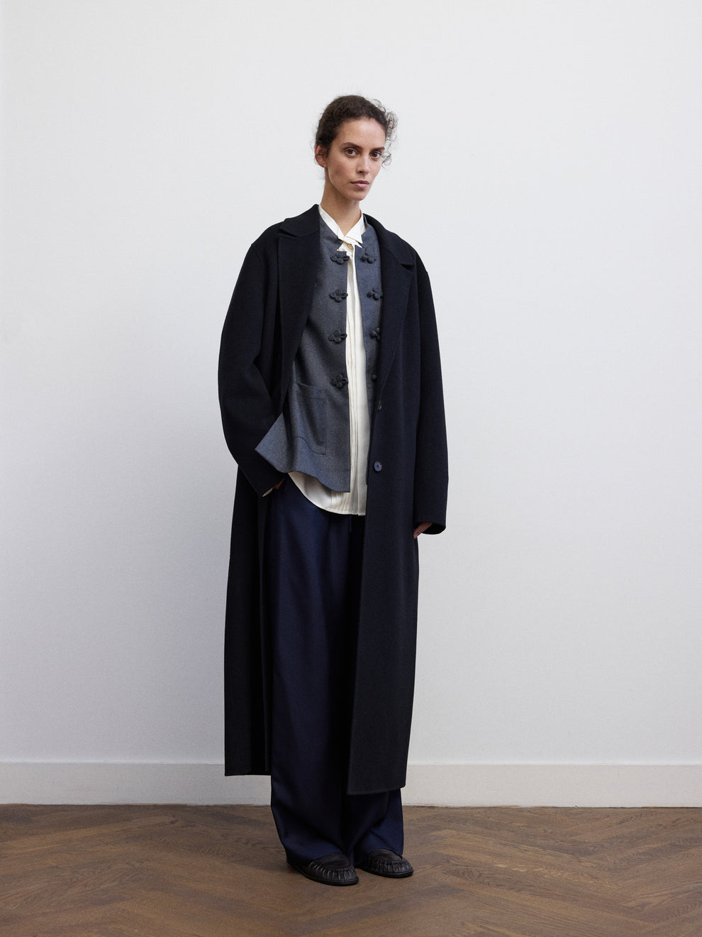 Woman wearing a black coat layered over a grey flanel jacket and navy silk pants against a white background