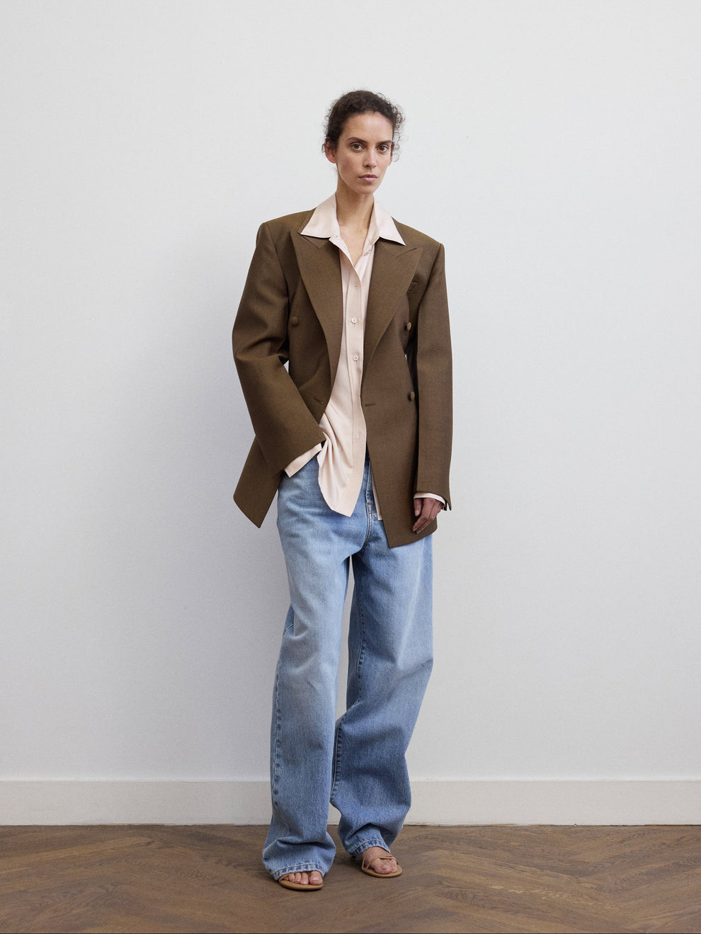 Woman wearing a light pink silk shirt with a golden brown colored blazer on top and jeans against a white background 