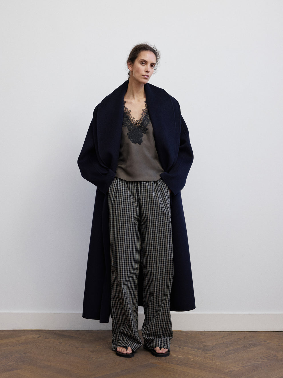 Woman wearing a lace v-neck silk top in an earth color, checked pants and a navy double faced wool coat against a white background