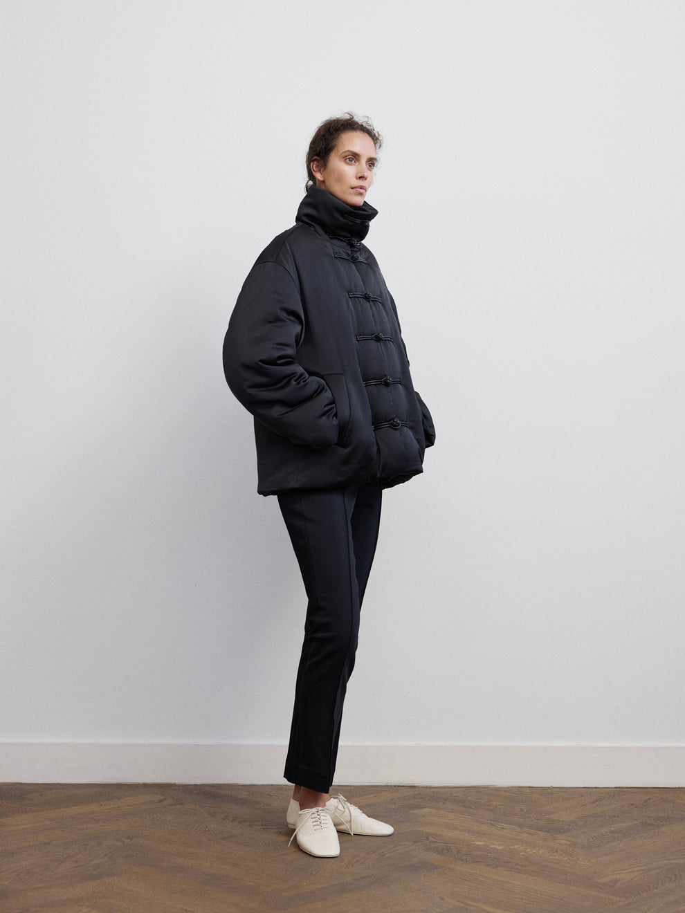 Woman wearing a black satin puffed jacket with mandarin closure and cigarette leg pants against a white background