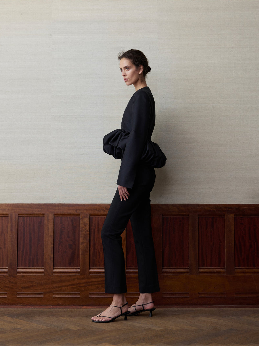 Woman wearing a long sleeve black top with a voluminous peplum bottom and cigarette leg trousers 
