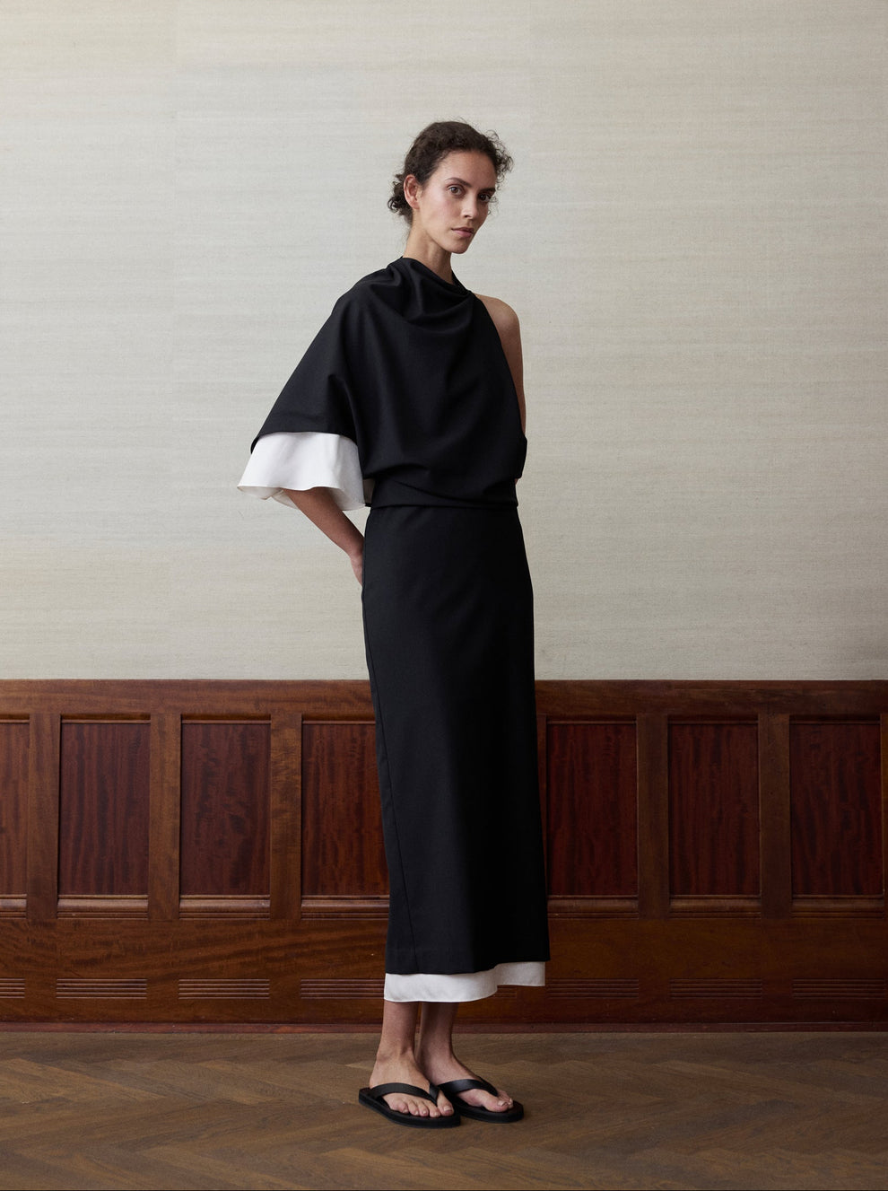 Woman wearing a one-shoulder black dress with a cream silk layer coming out from the bottom hem and left sleeve against a linen and wood paneled background