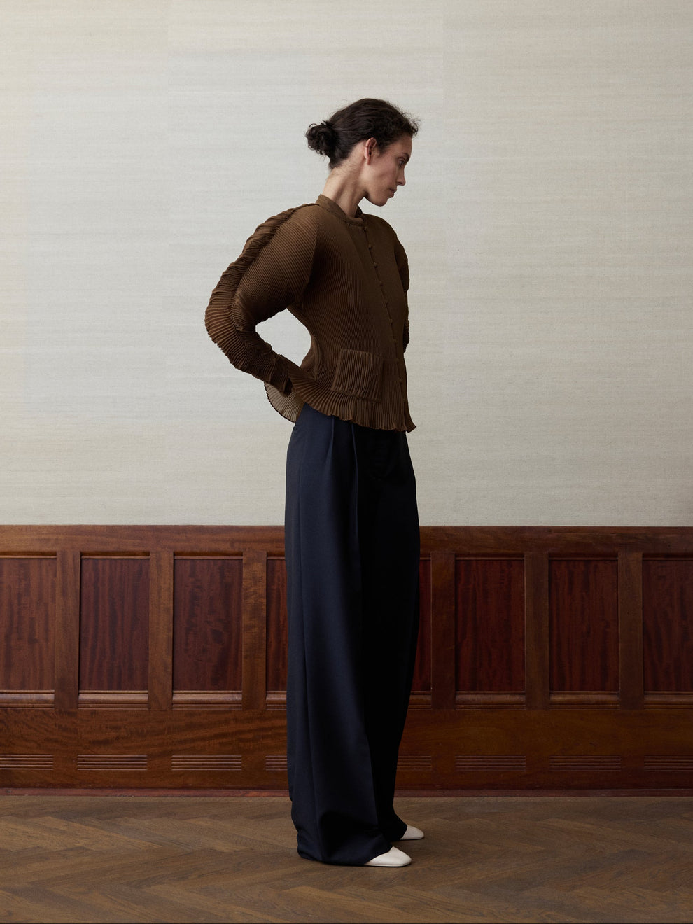Woman wearing a rust colored plisse cardigan and navy trousers against a linen and wood paneled background 