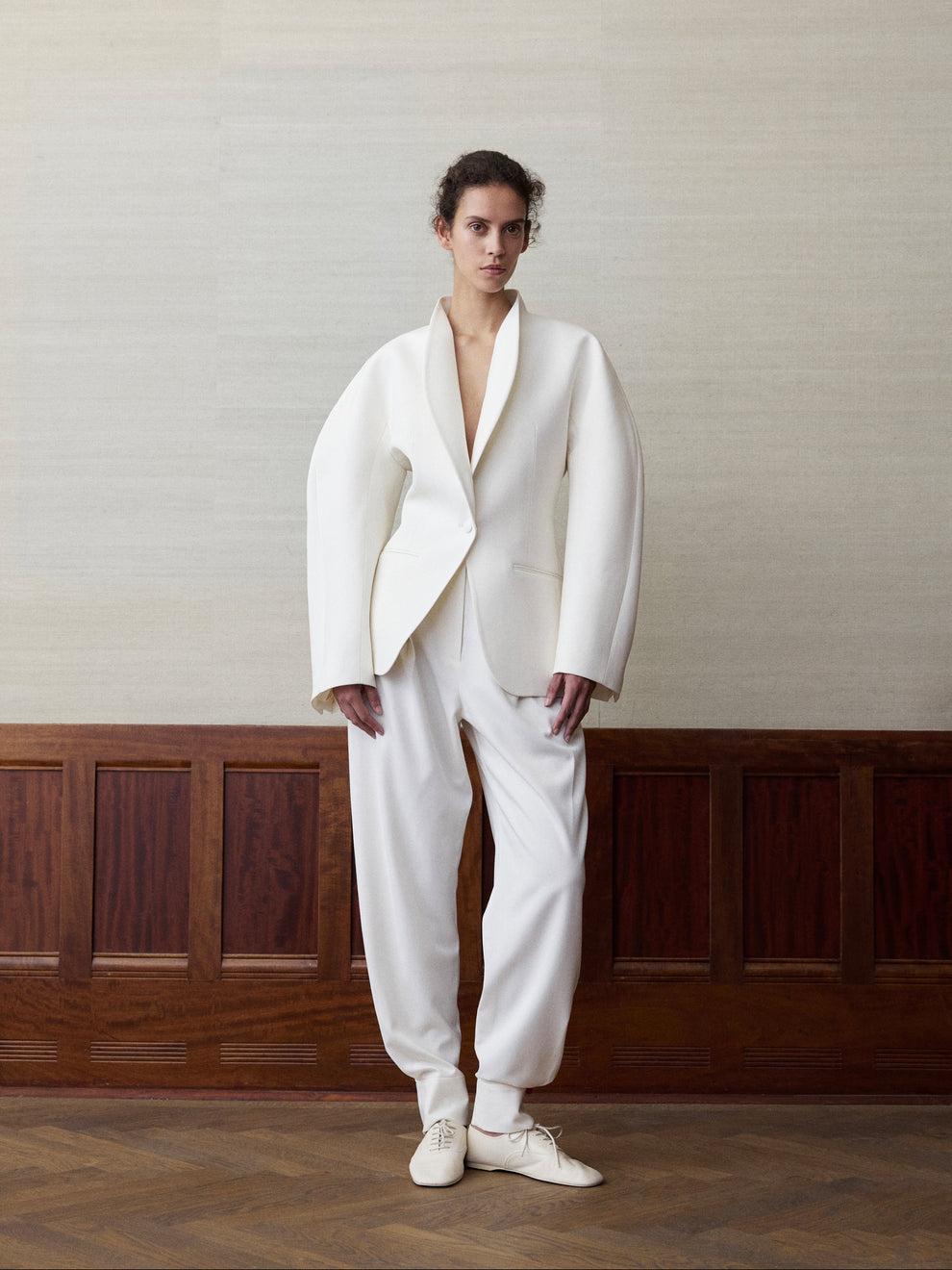 Woman wearing a sculptural offwhite blazer and offwhite balloon fit pants against a linen and wood paneled background