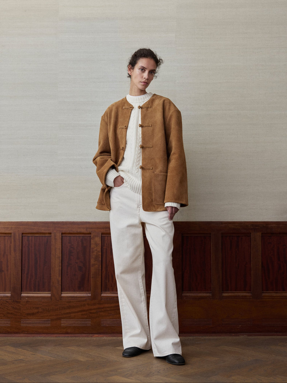 Woman wearing a offwhite cable knit sweater and suede cognac colored jacket and ecru denim against a linen and wood paneled background