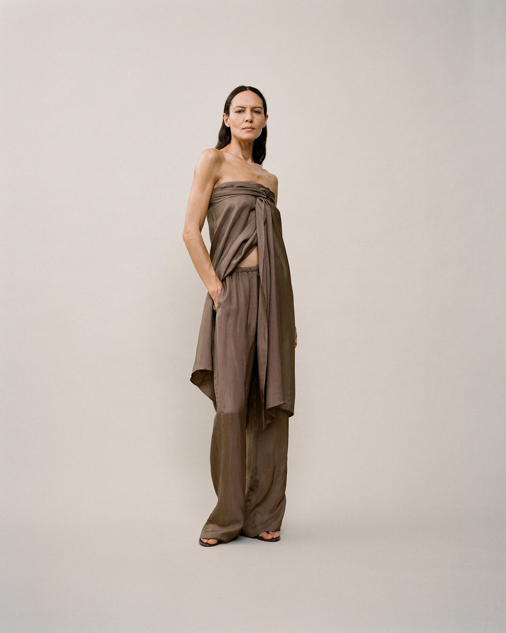 Person wearing a bron sarong top and brown trousers against a white backdrop 