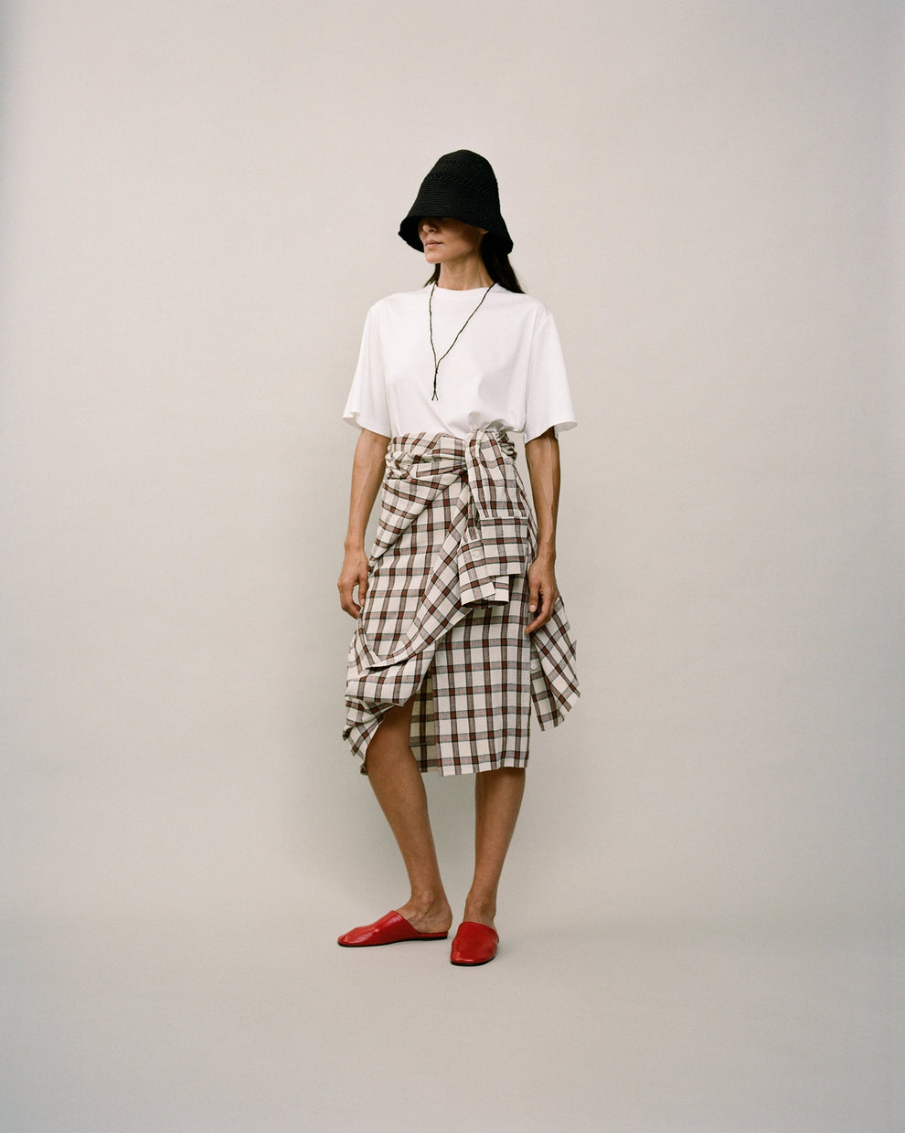 Person wearing a white t-shirt and a red checked wrap skirt against a white backdrop 