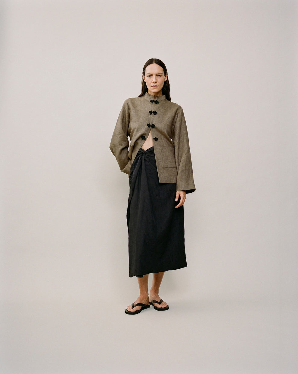 Person wearing a brown linen pankou closure jacket and a black sarong knot skirt against a white backdrop