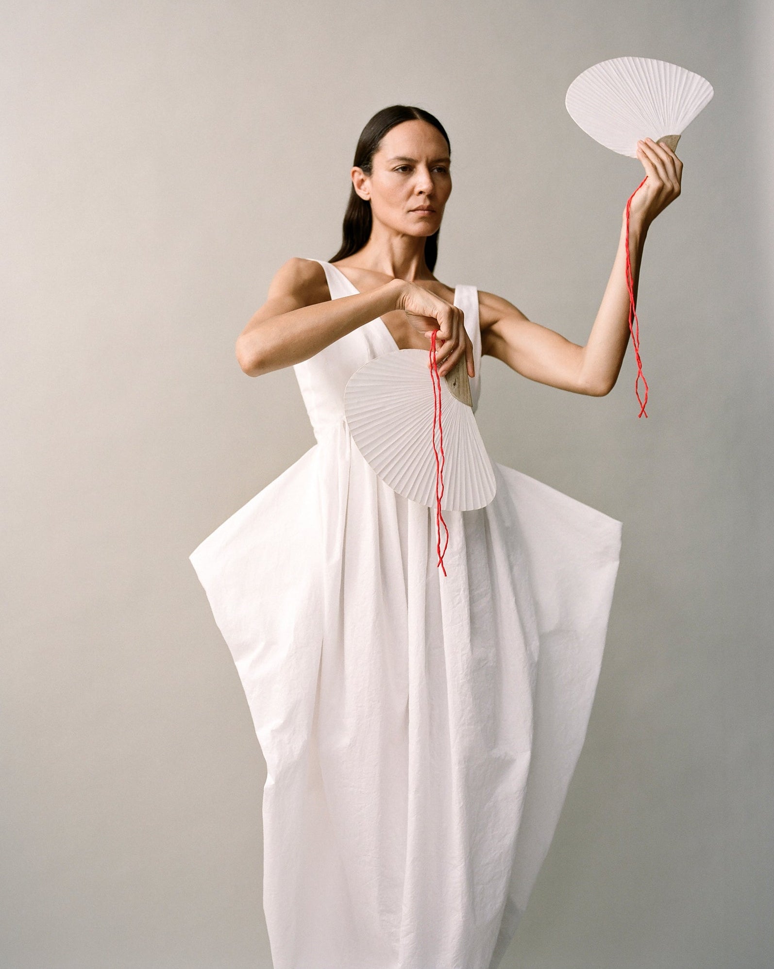 Woman wearing a white sculptural dress and two white fans with red strings. 