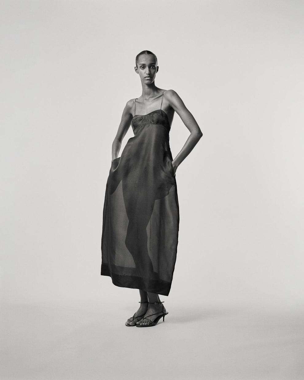 Black and white image of a person wearing a long sheer dress against a white backdrop 