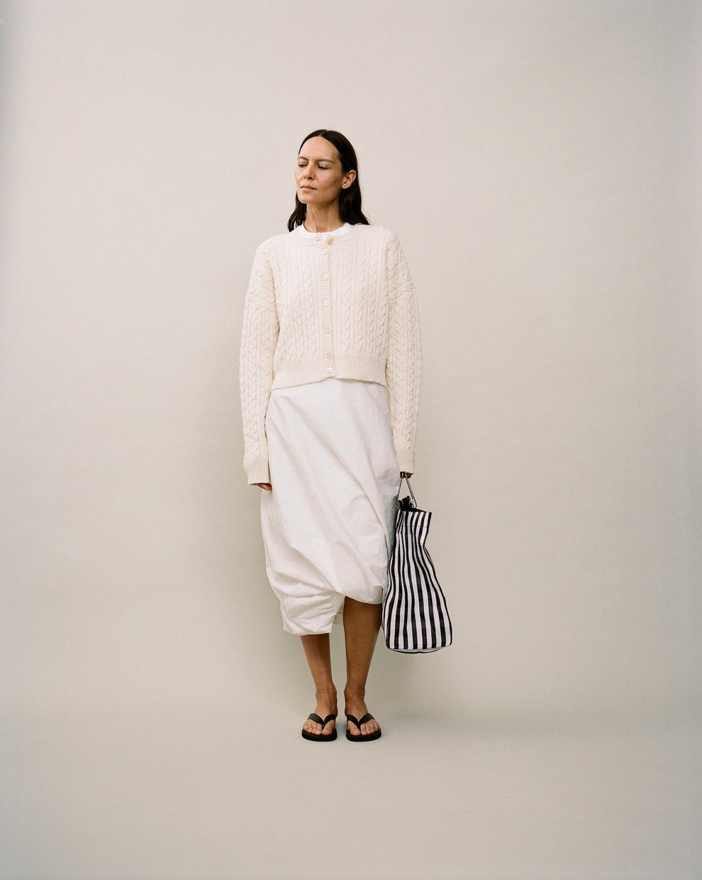 Person wearing a cream cable knit cardigan and a white twisted skirt against a white backdrop 