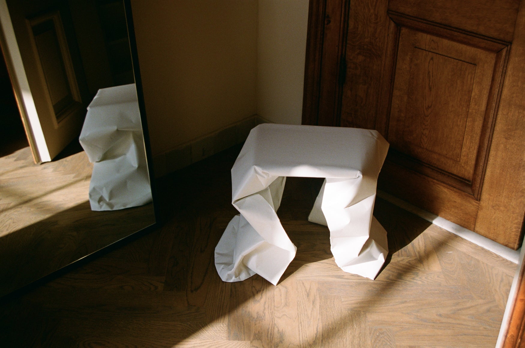 White stool looking like a piece of crinkled paper  in a room with wooden floor and door