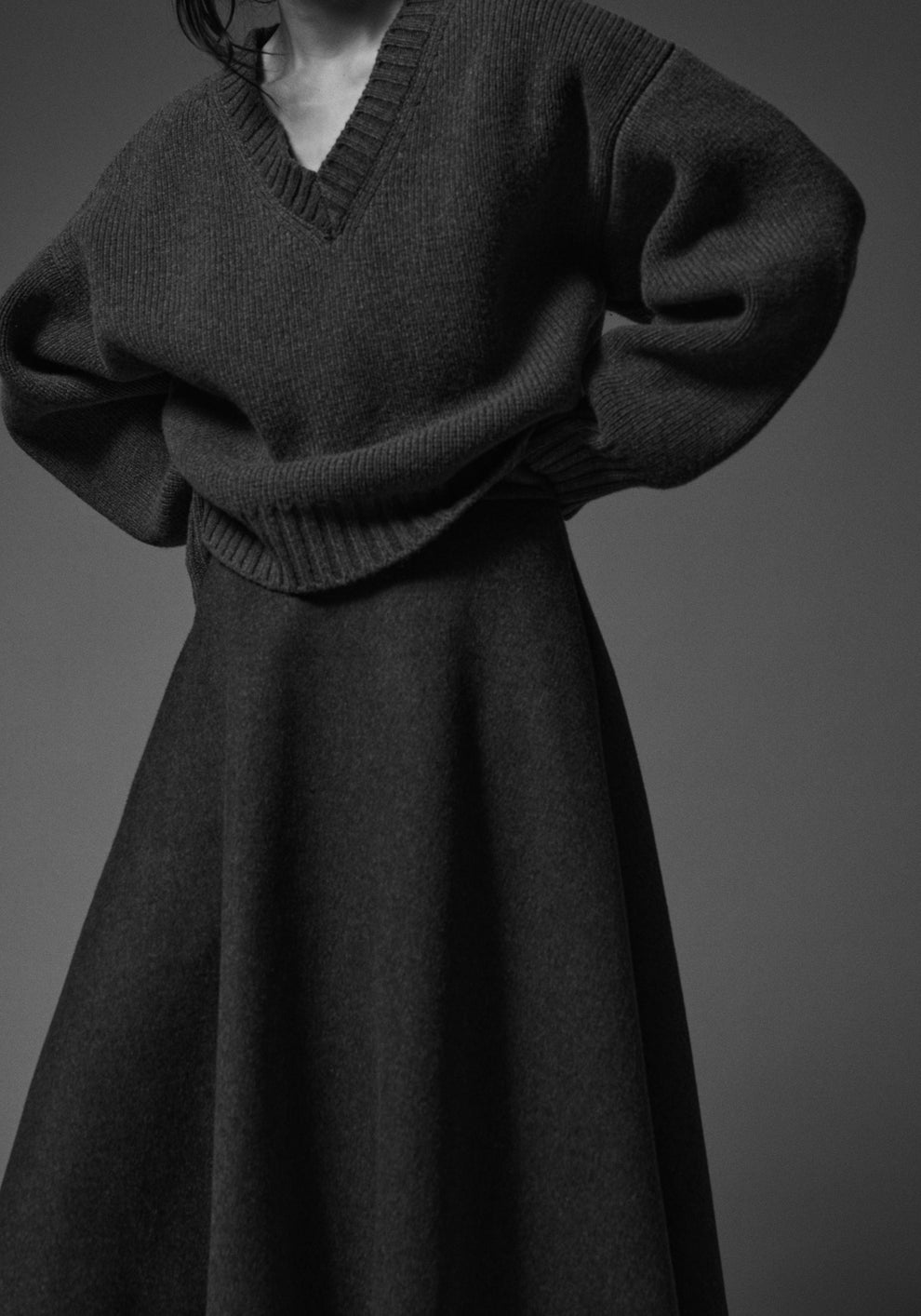 Person wearing a dark gray v-neck sweater and dark grey a-line skirt set against a plain background