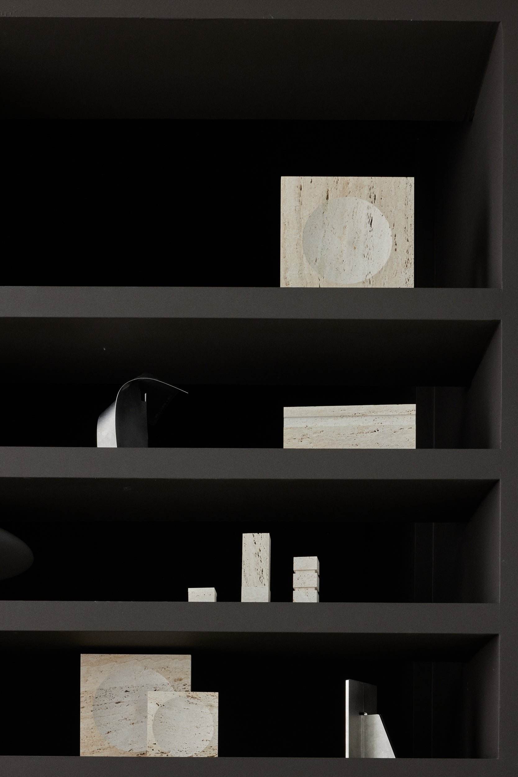 Dark shelving unit with various cylindrical travertin objects and a square item on a black background
